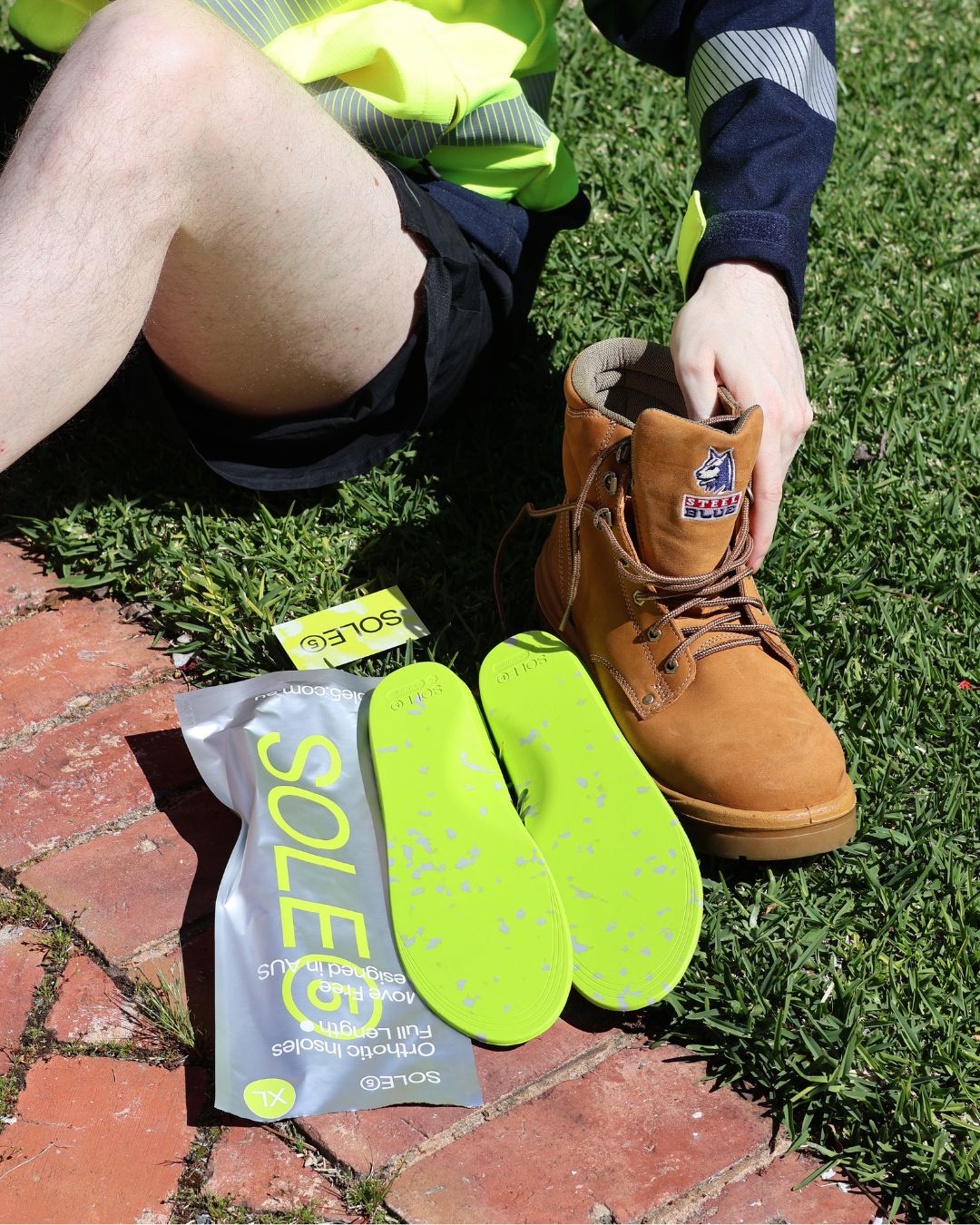 A person outdoors on grass and brick beside a brown work boot, with two bright neon green Sole5 cushioning inserts and a branded package laid on the ground. The person is wearing a high-visibility vest and shorts, and the boot has a visible Sole5 insole.