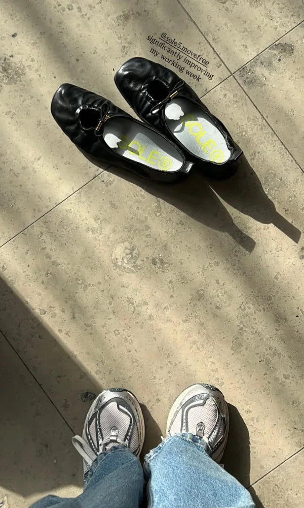 A top-down view of a person wearing white and gray sneakers with light blue jeans, standing on a sunlit concrete floor next to a pair of black leather shoes with SOLE5 Half Length orthotic insoles inside. The caption reads '@sole5.movefree significantly improving my working week'.