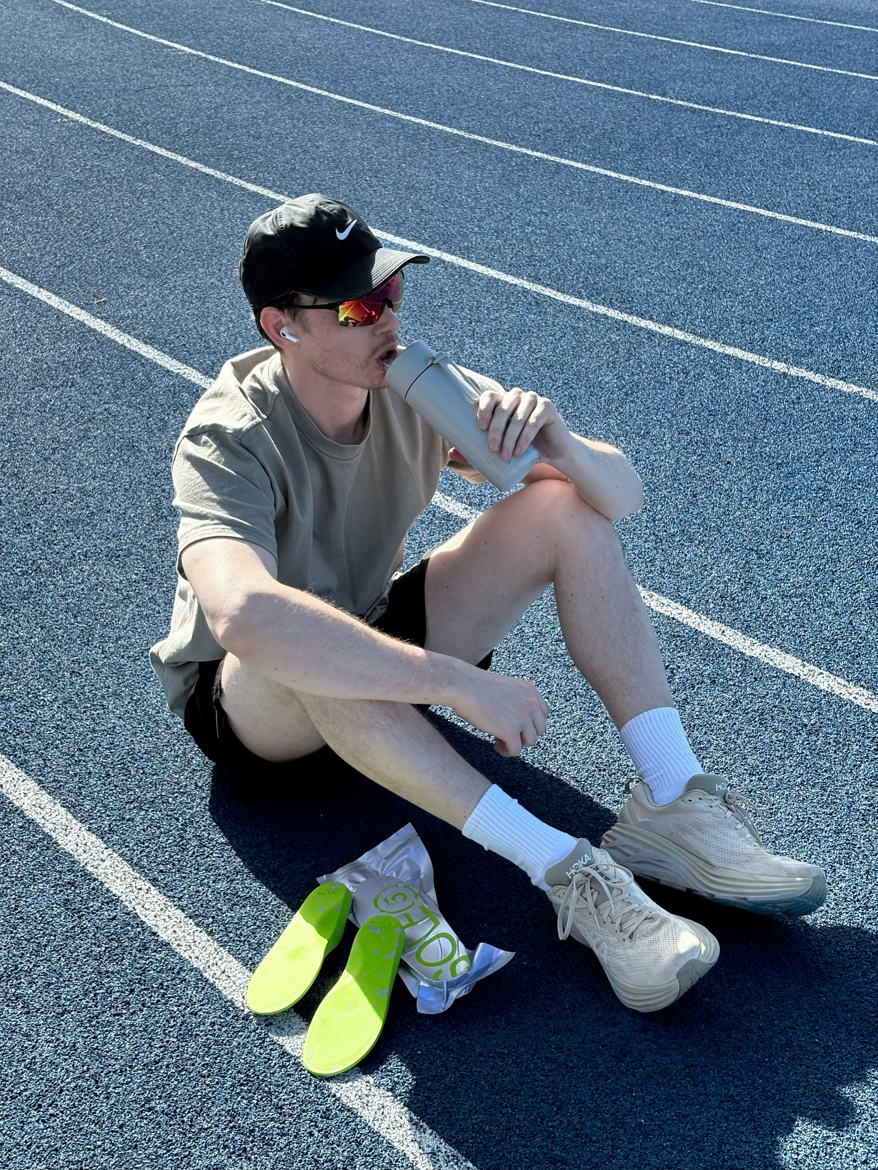 A man sits on a blue running track wearing a beige t-shirt, black shorts, white socks, and beige running shoes, holding a water bottle. A pair of bright green running shoes in their packaging lies on the track beside him.