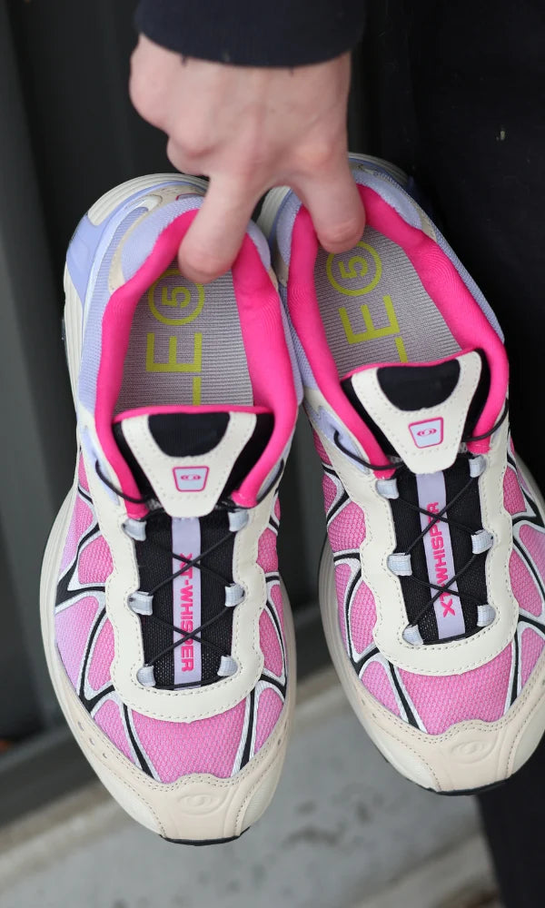 A pair of pink and white Salomon sneakers with black accents, held up by a hand against a dark background highlighting the SOLE5 orthotic insoles inside.
