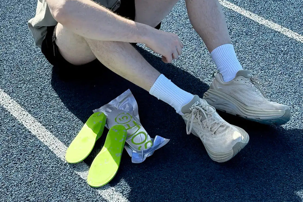 A man sitting on a blue running track drinking his water bottle. A pair of SOLE5 Orthotic Insoles are placed next to him