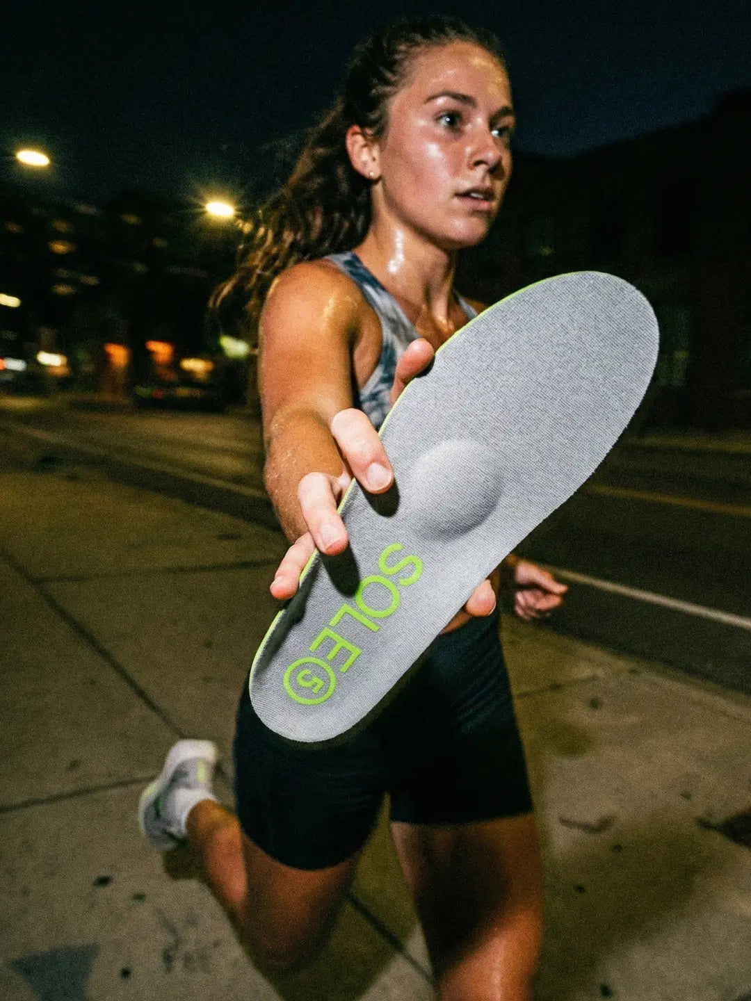 Woman running and holding a SOLE5 orthotic insole at the camera on a street at night