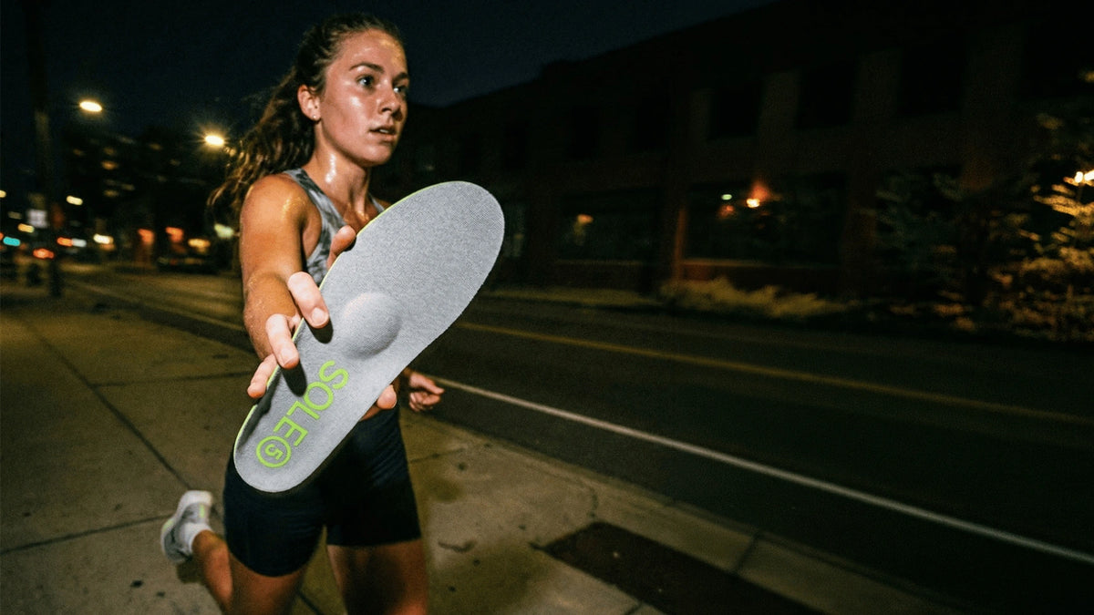 Woman running and holding a SOLE5 orthotic insole at the camera on a street at night